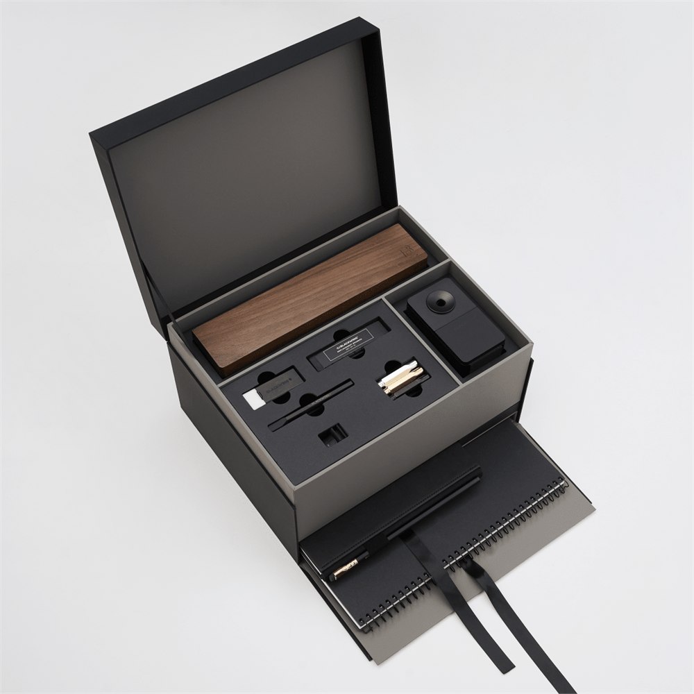 An open black gift box on a light gray background, revealing a wooden pencil box display set, blackwing handheld eraser, blackwing replacement erasers, blackwing point guards, and blackwing notebook clip on the top layer, and a large black spiral bound notebook and medium hardcover notebook with a black pencil in the holster in a lower pull-out drawer with a ribbon string.