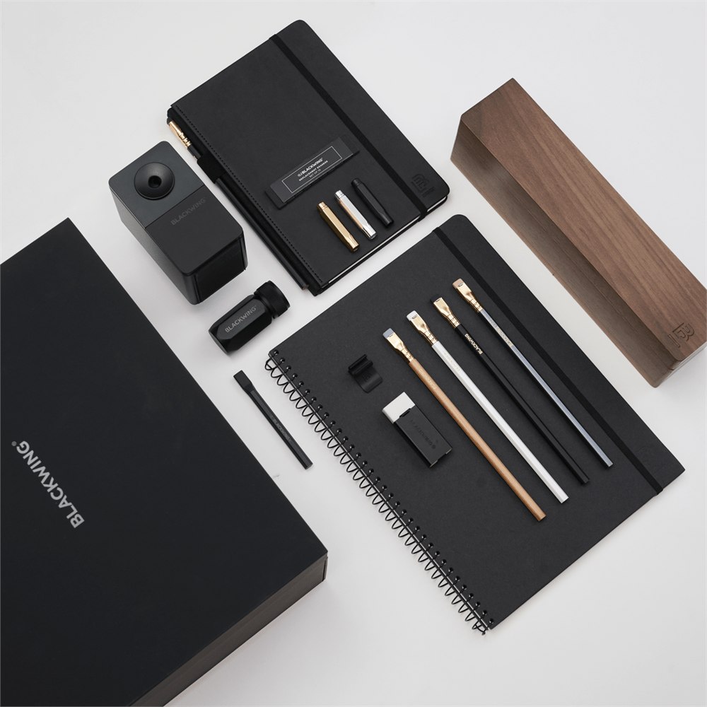 A black stationery and office accessory set arranged on a light gray background, including notebooks, a wood pencil display case, a pencil sharpener, a pencil extender, and other accessories and branded packaging. The visible brand text reads BLACKWING.
