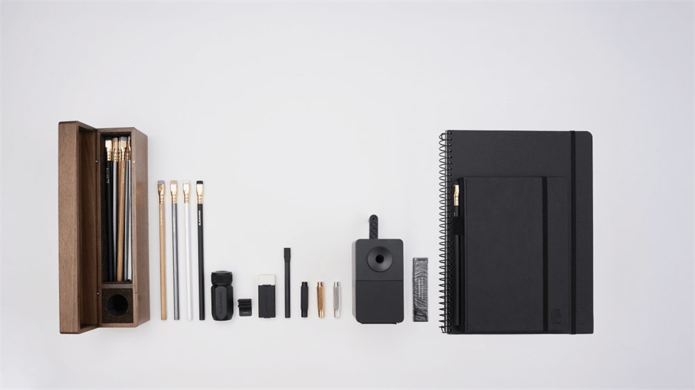 A flat-lay arrangement of stationery and art tools on a clean white background, including a wooden display case with pencils inside, a spiral notebook, a hardcover notebook, a black desktop sharpener, and small pencil accessories.