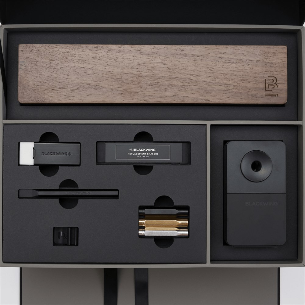 Blackwing stationery items set in a premium presentation box, featuring a wood display case, matte black pencil extender, black and white handheld eraser, black replacement erasers, a black desktop sharpener, a black notebook clip, and silver, gold, and black point guard pencil protectors.