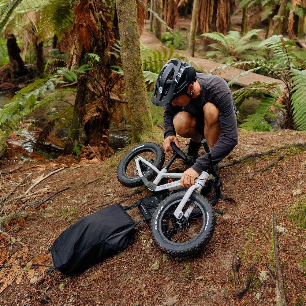 Adult loading a white balance bike onto the Kids Ride Shotgun Uplift backpack on a trail