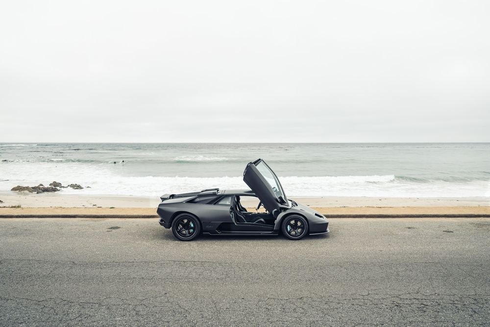 Side profile of the Eccentrica V12 Roadster with its door open by the coast