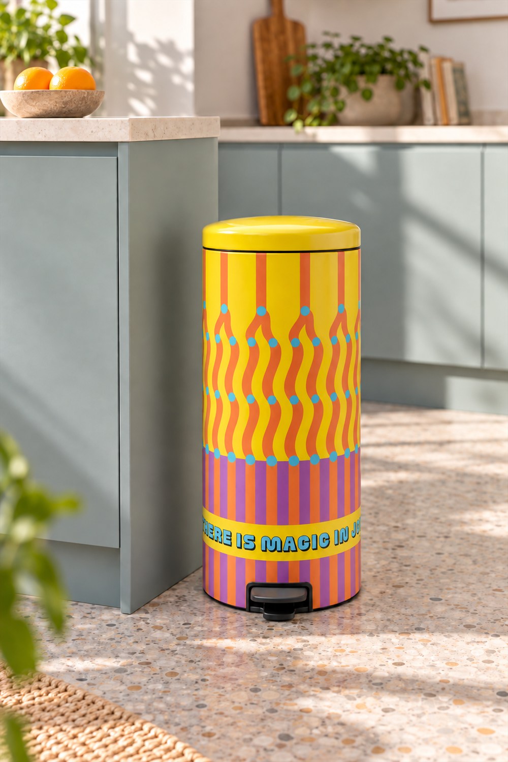 Tall yellow pedal bin with orange waves, turquoise dots, and purple stripes beside pale green kitchen cabinets on a sunlit terrazzo floor.