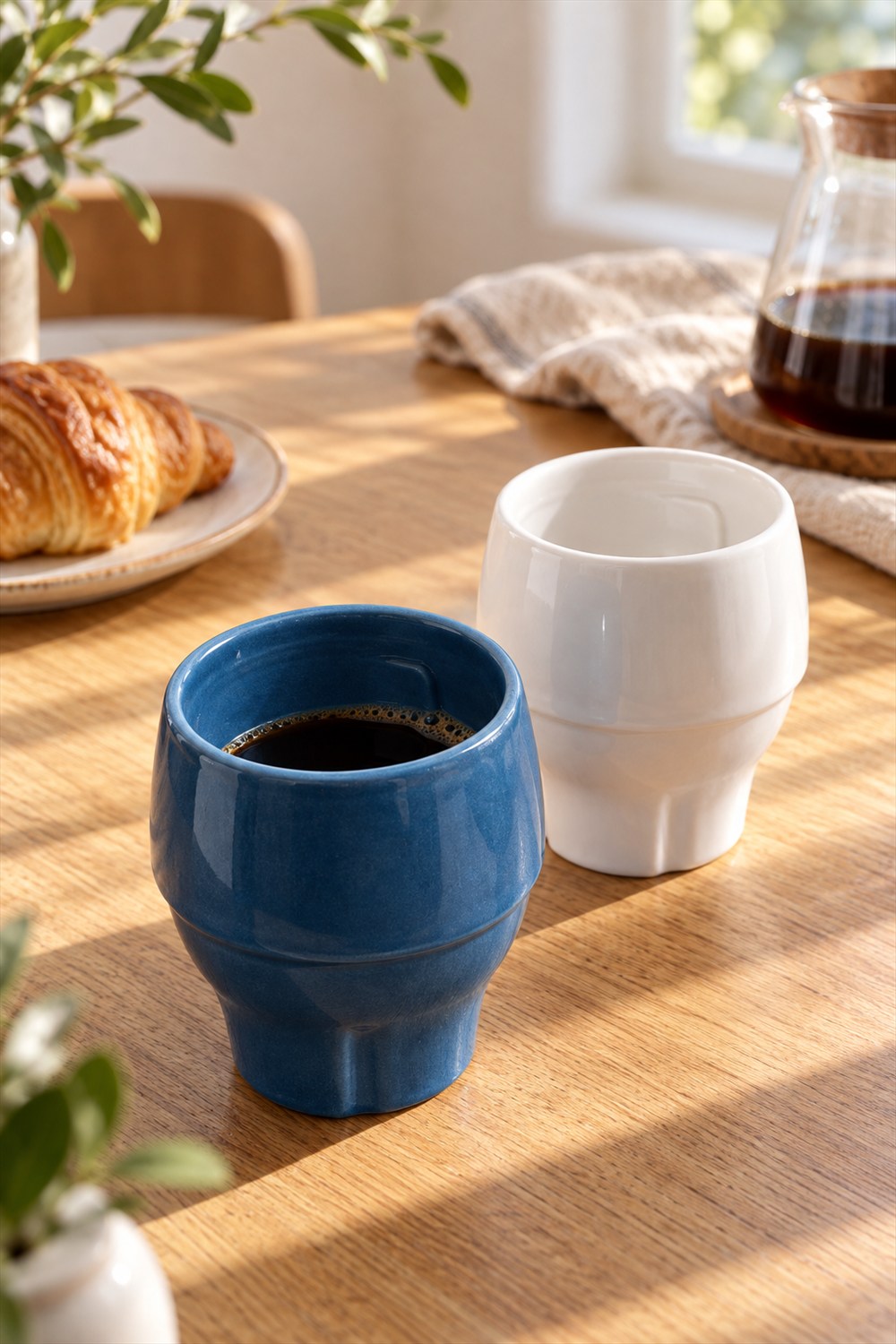 Blue and white handleless Avensi porcelain cups on a sunlit wooden breakfast table, with black coffee in the blue cup.
