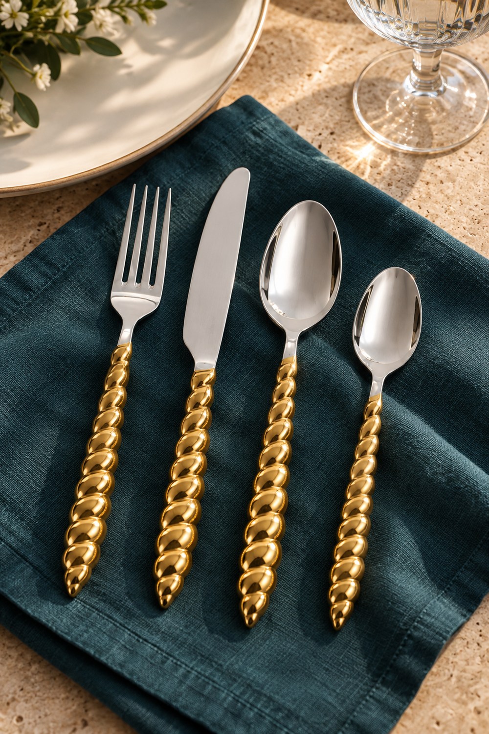 Four silver utensils with sculpted gold spiral shell handles arranged on a teal linen napkin beside a white plate and a glass.