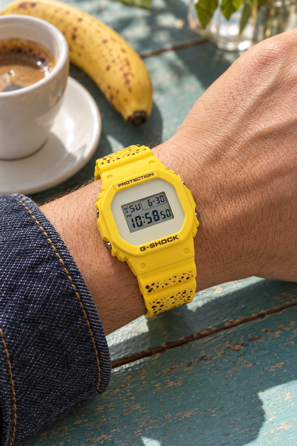 A bright yellow digital watch with brown banana freckles worn on a wrist beside an espresso and banana on a teal cafe table.