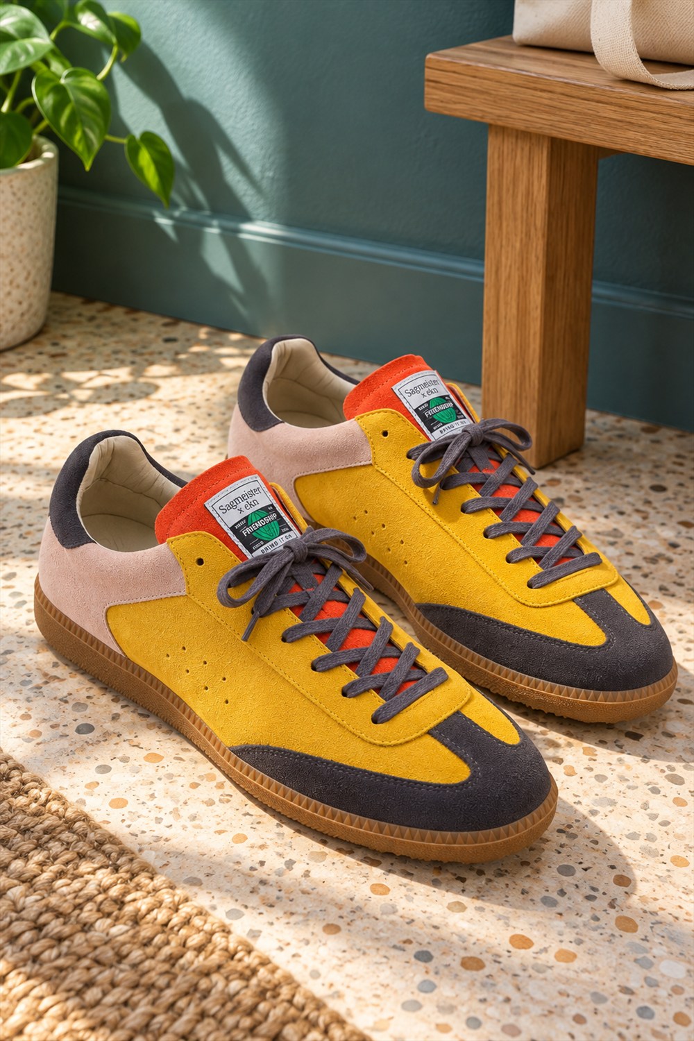 Yellow suede sneakers with charcoal toe panels, orange tongues, gray laces and pink heels on a sunlit terrazzo floor beside an oak bench.