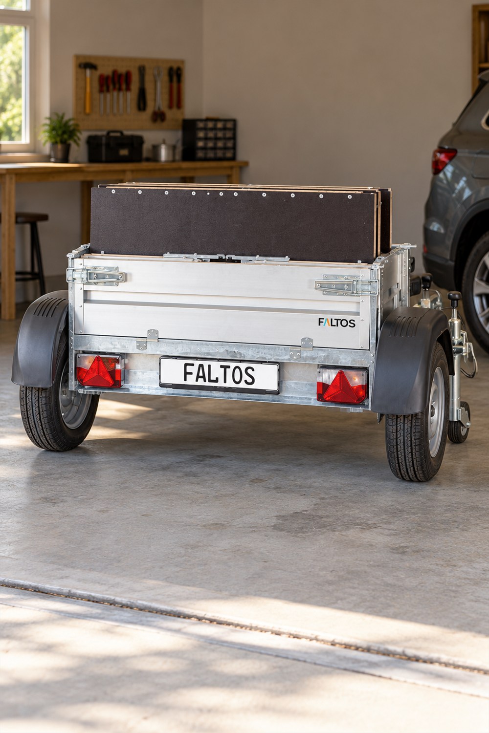 Silver FALTOS cargo trailer folded with dark floor panels upright between its wheels, parked on a concrete garage floor.