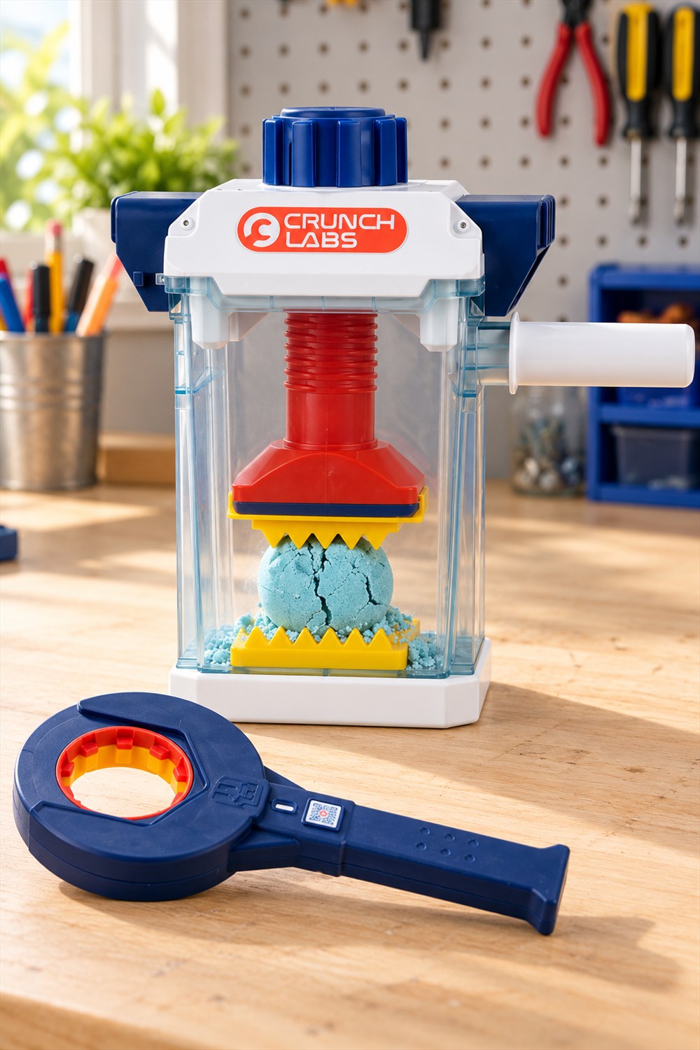 CrunchLabs Crunchinator with a red screw press and yellow jagged plates compressing a blue sand ball inside a clear chamber, with its navy wrench on a wooden table.