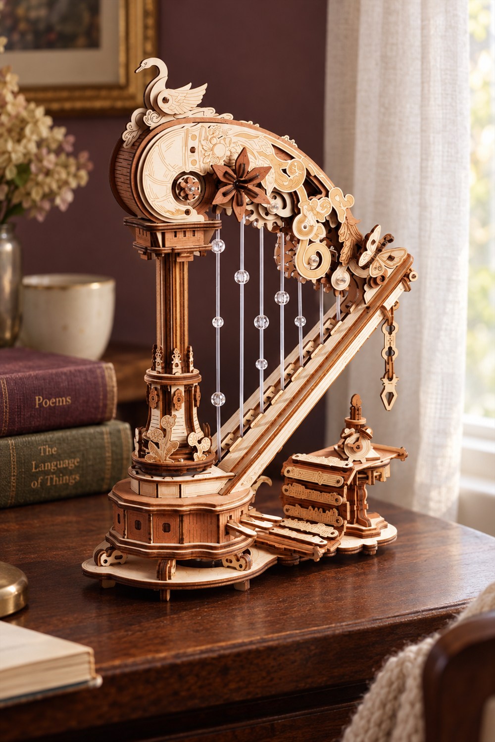 Wooden harp music box with exposed gears, a swan ornament, clear beaded strings, and layered base on a walnut table beside books and a sunlit window.