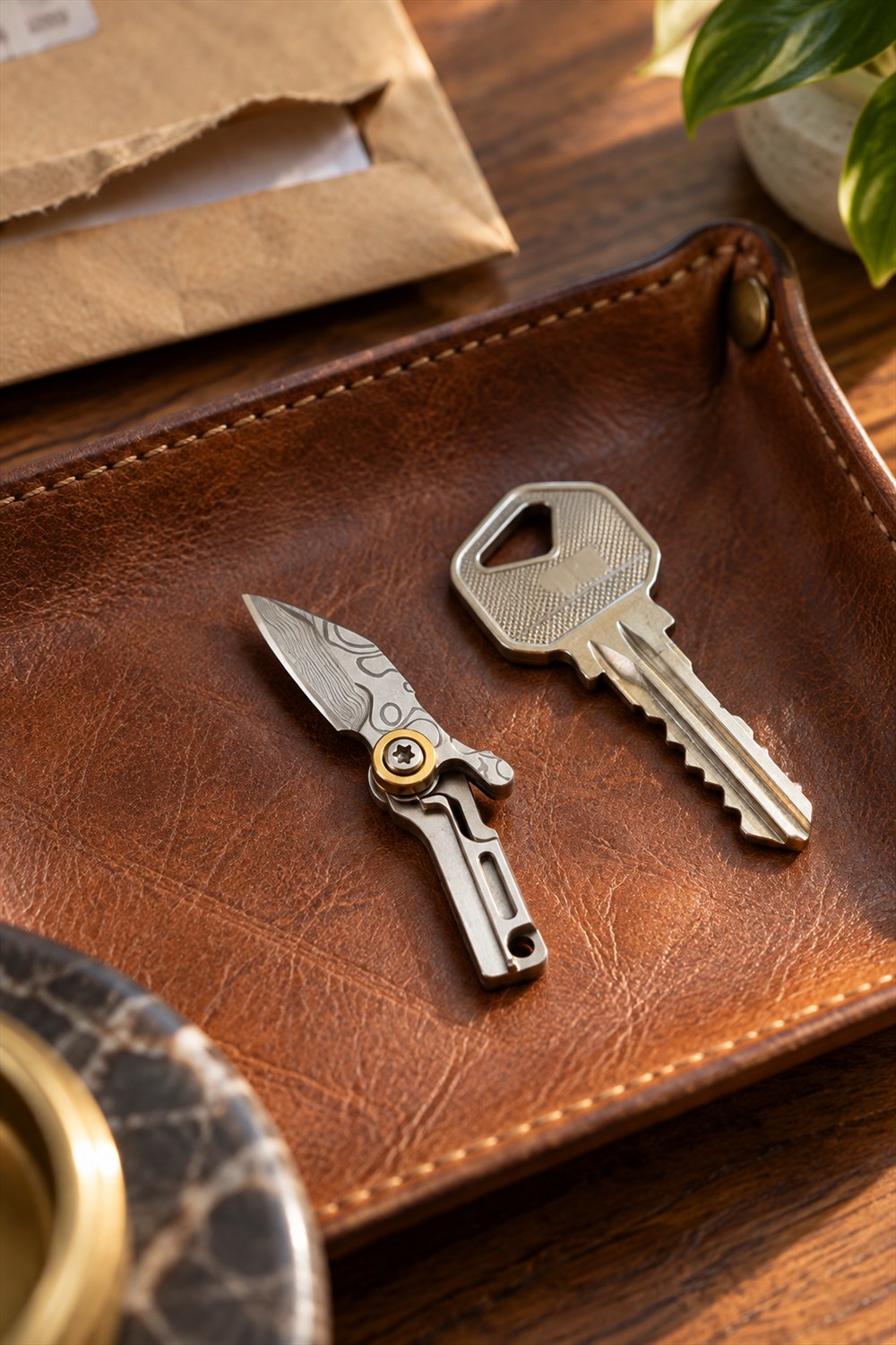 Open miniature folding knife with a patterned steel blade and silver handle beside a house key on a brown leather tray.