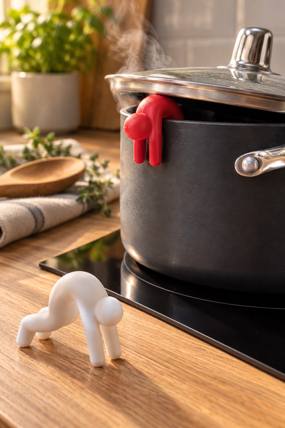 Red human-shaped silicone Lid Sid holding a steaming pot lid slightly open, with a matching white figure on the wooden kitchen counter.