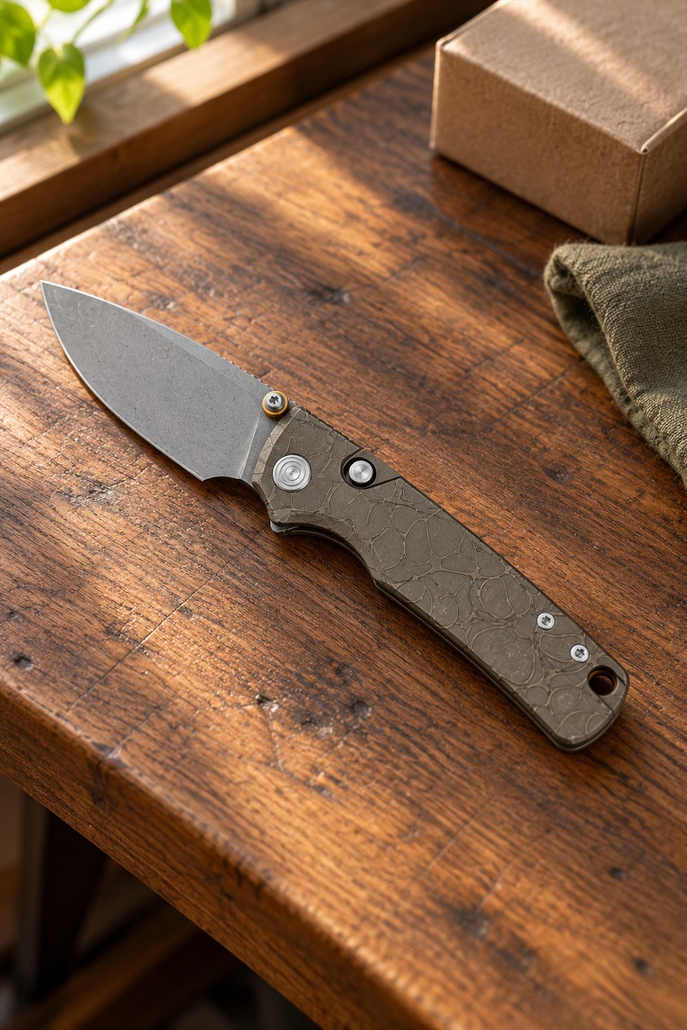 Open folding pocket knife with a gray blade and patterned titanium handle resting on a walnut workbench beside a kraft-paper parcel and olive cloth.