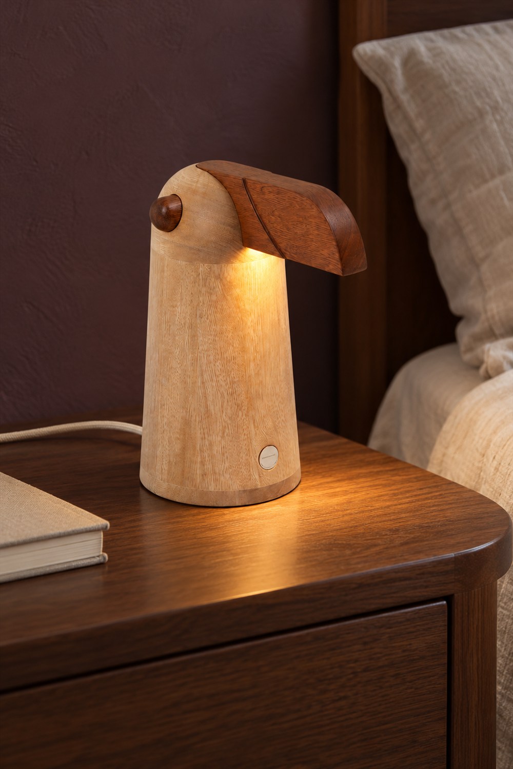 TUPI oak bird table lamp casting warm light beneath its dark wooden beak on a walnut bedside table beside cream linen bedding.